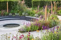 Compacted path spiralling around beds full of colourful summer perennials with inscribed wooden posts leading to central reflection pool. The Cancer Research UK Pledge Pathway to Progress - Hampton Court Flower Festival, 2019.