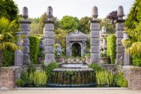 The Collector Earl's Garden at Arundel Castle, West Sussex in May. Designed by Isabel and Julian Bannermann. Green oak pilllars frame a grotto guarded by sea gods at the head of water representing the allegorical River Arun. Water runs over a shell lip before tumbling into a pool edged with Iris pseudacorus 'Variegata', ferns and Alchemilla mollis.