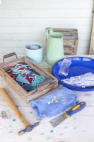 Smashed plates, mosaic tiles, powder grout, water, plate, scraper, hammer, mirror shards, cloth, gloves and goggles laid out on a wooden surface