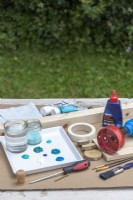 Paint, glue, wooden post, wooden board, wooden discs, paint brushes, paints, drill, cork, screwdriver, screws laid out on a table