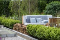 Outdoor seating next to a log pile and water rill, surounded by evergreen hedging - The Viking Friluftsliv Garden - RHS Hampton Court Festival 2021