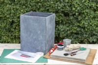 Large stone plant pot, stone slab, sash cord, masonry paint, sealant gun, adhesive, two paint brushes, craft knife, pencil, ruler, paper, masking tape, clear plastic sheet and a cutting mat laid out on a table