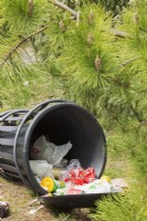 Tipped over garbage can with spilled recyclable paper and plastic drinking cups, packaging materials in a public park - April