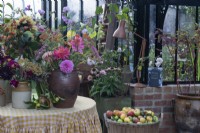 A table in the greenhouse that has a collection of vases of cut flowers, including: dahlias, sunflowers, cosmos,sedums and rose hips.