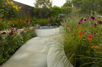 Raised herbaceous borders surrounding a paved seating area in the Finding Our Way An NHS Tribute Garden. Plants include: Dahlia,  Carex, Stipa giganta and Kniphofia.