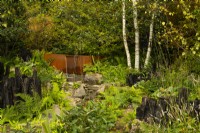 A woodland containing a fountain and stream running from a steel trough over rocks surrounded by trees, perennials and charred log borders in the Yeo Valley Organic Garden. 