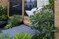 Hammock strung on wooden deck with slatted walls. Green plants in the foreground including foliage of oleander and Hedychium. Cyperus alternifolius next to water feature with white ginger lily, Hedychium coronarium behind. Calm of Bangkok.