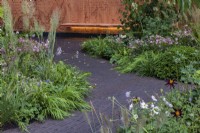 Carved cantilevred wooden bench with back lighting forms a focal point in a paved area with lush planting. Featuring Anemone x hybrida, Dahlia 'Verrone's Obsidian', Astrantia, Hakonechloa macra, and Pennisetum alopecuroides cvs. The Florence Nightingale Garden.