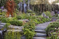 Stone steps and path through the garden with metal sculpture and a waterfall. Planting including Salvia nemorosa, Persicaria amplexicaulis, Veronica longifolia, Astrantia major, Geum coccineum 'Cooky', chamomile, pine, ferns and Achillea millefolium. Bodmin Jail: 60Â° East - A Garden Between Continents, RHS Chelsea Flower Show 2021 