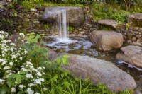 Psalm 23 Garden. Naturalistic cascade of water from granite, worn rocks in drystone wall into tranquil pool. Featuring Eupatorium 'Lucky Melody' and young shoots of Viburnum opulus .