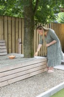 Special lighting. Large wooden lounge bench with woman under the tree. Path of gravel. Wooden fence.