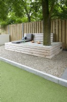 Large wooden lounge bench with cushions and candles under the tree. Path of gravel. Wooden fence.