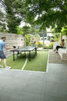 Man and woman playing at the pingpong table. Large wooden lounge bench with woman under the tree near the pingpong field.