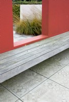 Wooden bench attached to a red wall with view through to ornamental grasses, patterned tiles in foreground.