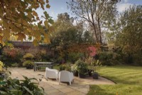 Stone patio area with metal table and modern chairs looking out onto garden with groups of pots, borders, mature shrubs and trees - October