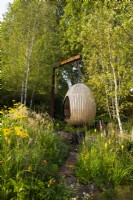 A path leading to egg shape swing seat surrounded by Kniphofia 'Tawny King', Rudbeckia laciniata 'Herbstonne', Calamagrostis acutiflora 'Karl Foerster', Calamagrostis brachytricha. The Yeo Valley Organic Garden. Designer: Tom Massey, supported by Sarah Mead, Chelsea Flower Show 2021. 