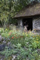 The Blue Diamond Forge Garden. Thatched hut with stone walls. Blacksmith working on anvil inside. Planting includes Crab-apple Malus 'Wintergold', Crocosmia 'George Davison' Montbretia, Helenium 'September Gold', echinacea, coneflower, Melica altissima, kniphofia and Helenium 'Kleine Aprikose'. Designers: The Blue Diamond Team. 