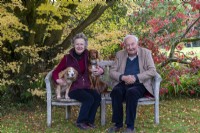 Lawrence and Elizabeth Banks sit with their dogs on a bench beneath maples.