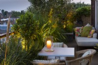 At night, a dining table on a balcony rests beside planters with a standard bay tree, rosemary, phormium and palm.