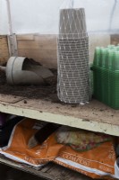 A bulk load of green plastic seed trays and grey plant pots sit on a potting bench beside a pile of compost and a wooden handled scoop in a small commercial nursery with a bag of compost below. Spring.