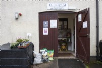 An entrance to a small nursery with some plants on a stall to the left of the entrance door, bags of compost and other sundries beside the door and primroses, log fuel and fish, blood and bone inside the entrance. Spring. 