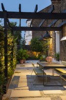 Illuminated dining area in long narrow space with table and chairs under pergola with fitted patio heaters. 