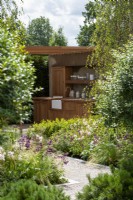 View of outdoor kitchen through planting of Gaura, Diascia and grasses surrounded by birch trees and Osmanthus x burkwoodii shrubs.  The Viking Friluftsliv Garden, RHS Hampton Court Palace Garden Festival 2021