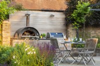 View across beds to outdoor dining area with wooden table, chairs and bespoke kitchen unit with integrated pizza oven and bbq grill. 