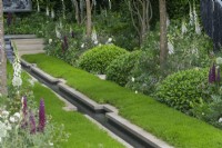 A central rill runs through camomile lawns edged in borders of white foxgloves, alliums, peonies, with pink lupins.