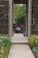 Woven willow panels enclose a doorway that frames the view of a bronze urn beyond. A straight, paved path is edged in golden aquilegia and grasses.