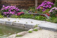 Bespoke iron bench overlooking a pond with colourful Rhododendrons behind - A Swiss Sanctuary, RHS Chelsea Flower Show 2022