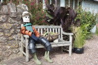 An English village flowershow competition display featuring a scarecrow flowerpot man sitting on a bench next to a basket of homegrown vegetables.