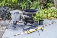 Bucket of slate, faux barrel container, ferns, drill, saw, trowel, copper pipe, silicon, boat level, water pump, pen, zip ties, pipe cutter, wire wool, metal grill laid out on the ground