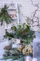 Winter evergreen foliage including Ilex and conifer -displayed in glass jars in metal bottle carrier and on table with tea lights