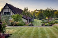 Looking down striped lawn and borders next to gabled house at sunrise. Plants include a pair of Tibetan cherry trees (Prunus serrula).