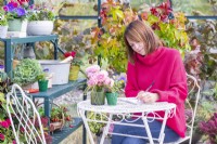 Woman sitting at a table writing in a greenhouse that has been filled with various plants and mixed containers