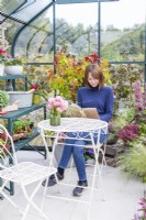 Woman sitting at a table reading in a greenhouse that has been filled with various plants and mixed containers