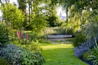 View along grass path, with border either side, to a boat moored on a pond.