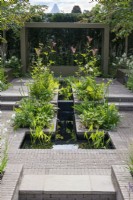 A paved, terraced garden has a central rill carrying water from an upper to a lower pond. Along its margins are white and green themed borders of ferns and agapanthus.