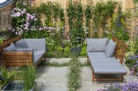 A small relaxing courtyard has fleabane growing between cracks in the paving, borders of daisy like flowers, and roses or fruit trees trained up wooden walls.