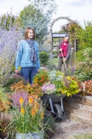 Woman pushing wheelbarrow full of plants through garden with woman sweeping in background