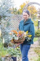 Woman holding an arrangement of Cotinus 'Grace', Eucalyptus 'Silver Dollar', Eryngium 'Picos Blue', Anemone 'Ruffled Swan' and Astrantia 'Snow Star' in a wooden trug