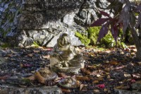 A buddha ornament sits at the base of a retaining wall beside an acer tree. Whitstone Farm, Devon NGS garden, autumn