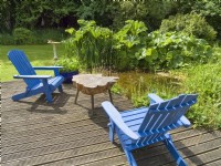 Painted wooden adirondack chairs and rustic table on decking overlooking natural garden pond