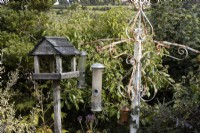 A white, rusty bird feeding station with various feeders stands beside an old wooden bird table. Autumn.