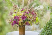 Chrysanthemum 'Tula' mixture seed heads of miscanthus and sprays of rose hips November. Cut flower arrangement in a vintage pottery jar on a garden table.