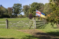 A gravel drive leads to a post and rail wooden gate and fence leading into a large grassy field. A Union Jack and Ukraine flag flies from the fence. Regency House, Devon NGS garden. Autumn