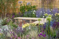 Insect hotel built into a wood and gabion bench surrounded by planting of Salvia 'Purple Rain', Gaura, Stipa tenuissima, Agapanthus 'White Heaven', Perovskia 'Blue Spire' and Achillea 'Credo' - Turfed Out Garden, RHS Hampton Court Palace Garden Festival 2022