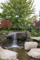 Natural waterfall over a large stone boulder into a pool, bright orange leaves and fruits of Viburnum opulus behind - Bible Society: The Psalm 23 Garden, RHS Chelsea Flower Show 2021