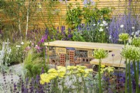 Bug or insect hotel inside gabion bench with wooden  top in low maintenance gravel garden with drought tolerant plants to attract pollinators, such as Salvia, Achillea, Agapanthus,Perovskia,  Stachys byzantina and Stipa tenuissima- Turfed Out, RHS Hampton Court Palace Garden Festival 202